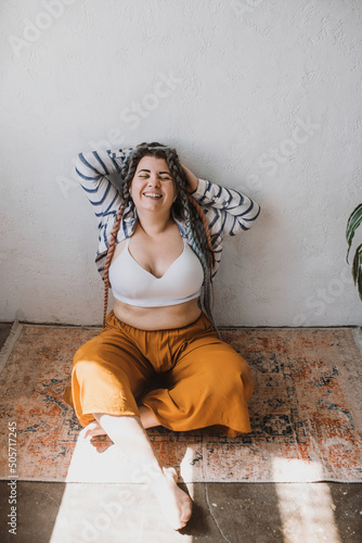 Positive smiling plus size woman with dreadlocks in bright yellow pants in a modern bohemian boho interior is meditating surrounded by large flowers and plants. Ecological approach to conscious consum photo