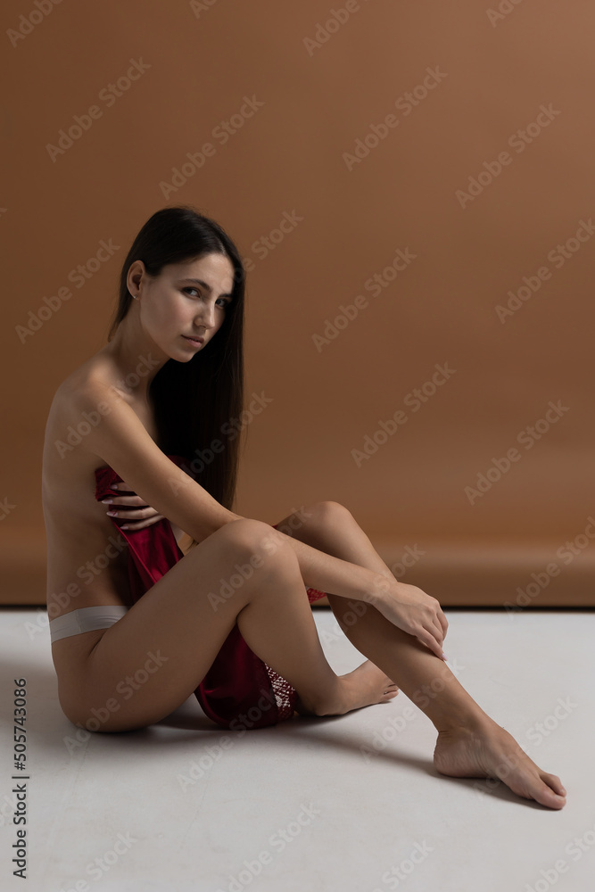 young and slim brunette European girl sensually poses topless in her underwear holding a red dress. Covering her breasts with the fabric of the dress on a brown ochre background sitting on the floor young and slim brunette European girl sensually poses topless in her underwear holding a red dress. Covering her breasts with the fabric of the dress on a brown ochre background sitting on the floor