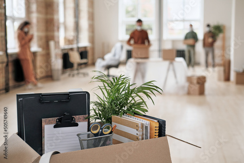 Focus on open cardboard box with office supplies, clipboards and plant against people moving into new office in background