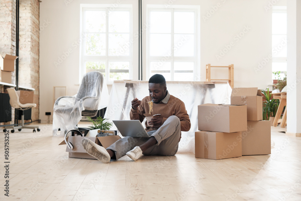 Too busy young black manager sitting with laptop on floor and eating noodles at workplace to finish project on deadline in office with moving boxes Too busy young black manager sitting with laptop on floor and eating noodles at workplace to finish project on deadline in office with moving boxes