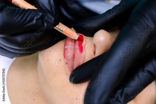 close-up of a permanent makeup master holding a red pencil and painting over the model's lips to perform a tattoo