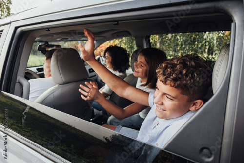 Teenage Boy and Girl Greeting to Someone Through Window with Waving Hands While Sitting Inside Minivan Car, Happy Four Members Family on a Weekend Road Trip photo