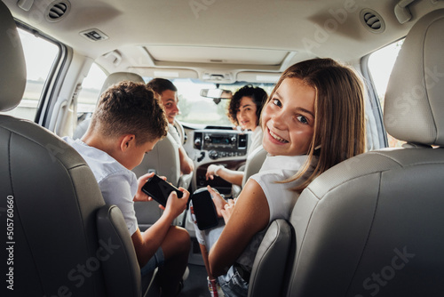 Cheerful Caucasian Teenage Girl Smiles Into the Camera While Sitting in Minivan Car with Her Brother, Mother and Father, Happy Four Members Family Enjoying Weekend Road Trip photo