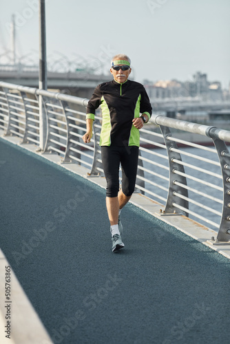 Vertical shot of modern strong senior man wearing tracksuit jogging along track in the morning