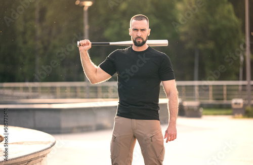 Sexy, strong, young man with a beard outside holding a bat on his shoulder
