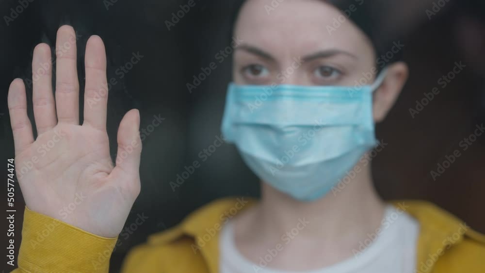 Close-up female hand on glass with blurred sad woman on coronavirus face mask at background. Upset Caucasian young ill lady standing indoors on Covid-19 pandemic quarantine looking out window Close-up female hand on glass with blurred sad woman on coronavirus face mask at background. Upset Caucasian young ill lady standing indoors on Covid-19 pandemic quarantine looking out window