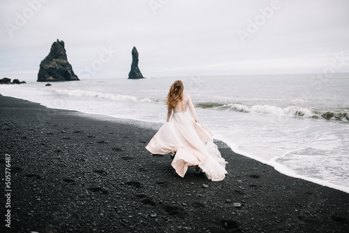 Girl in old old-styled wedding dress are running on the Black Sand beach in Vic, Iceland with rock islands on background.
