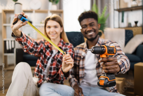 Blur background of multi ethnic couple in casual clothes showing measure tape and electric drill on camera. Cheerful family using tools for repairing and assembling furniture at new house.