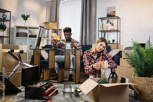 Positive woman unpacking boxes and talking on mobile phone while man assembling furniture behind. Young multiracial couple enjoying moving process to new house. photo