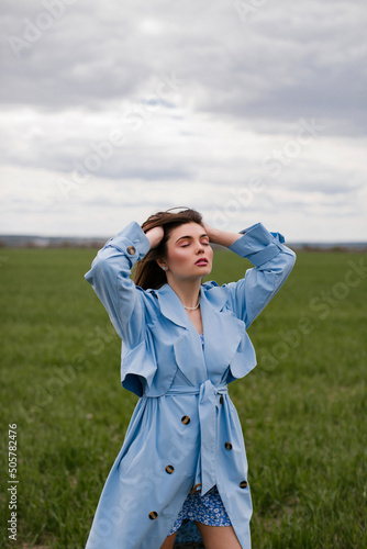 Stylish young woman with makeup and long dark hair posing in a blue trench coat in a green field.