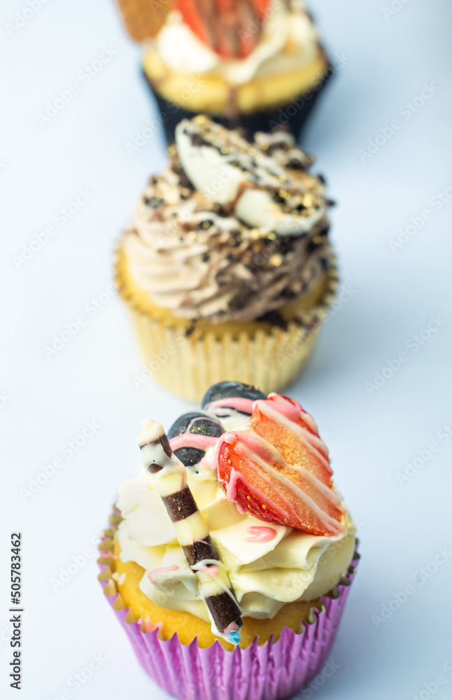 Beautiful muffin cupcakes decorated with fruits, chocolate, golden sprinkles, and cookies, in one line. Many pastries on light background.