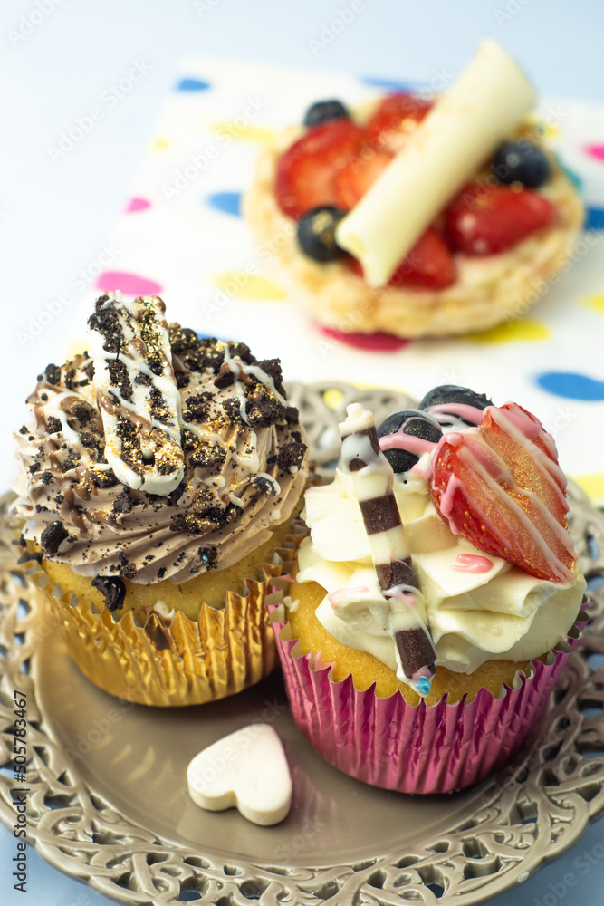 Two delicious muffin cupcakes decorated with cream, fruits, and chocolate on a romantic openwork plate with a candy heart. Little fruit tart and spotted napkin in the background.