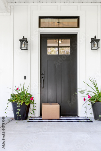 A blank cardboard box package delivered to the front door of a modern farmhouse style home. A package box shipped from e-commerce online shopping. © LUGOSTOCK