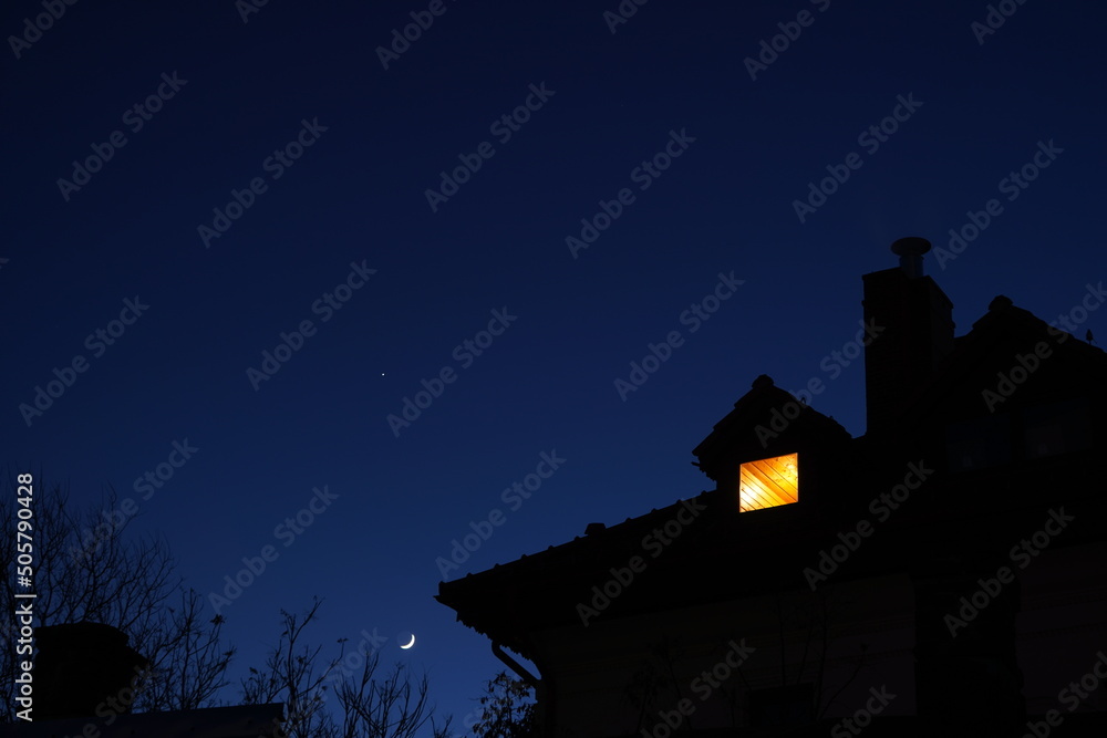 Φωτογραφία Silhouette of house roof with chimney and lit attic small window at night agains
