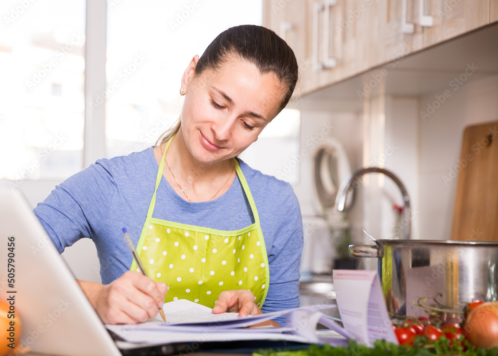 Portrait of woman filling papers and using laptop at kitchen
