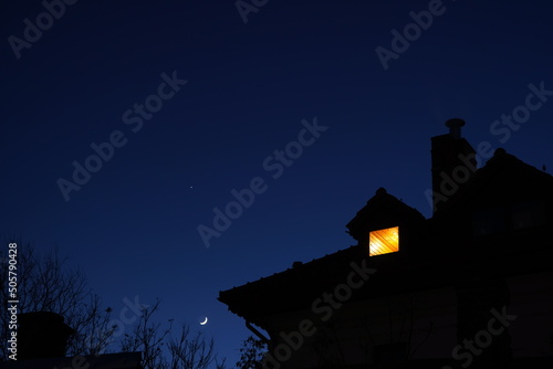 Silhouette of house roof with chimney and lit attic small window at night against clear sky with moon and star photo