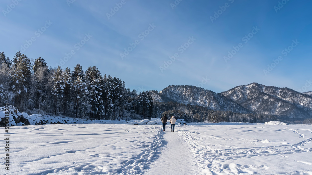 Fototapeta premium Two people walk away along a path trodden in the snow. Ahead, against the blue sky - a mountain range, a coniferous forest. Footprints in snowdrifts. Altai