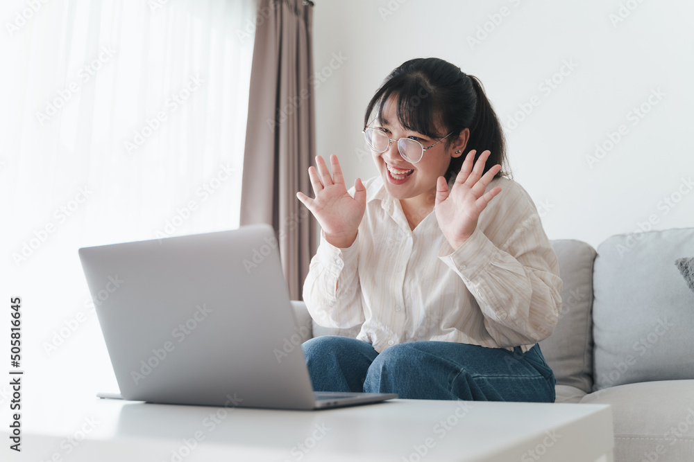 Young Asian woman using laptop computer for online video conference call waving hand making hello gesture on the couch in living room. Young Asian woman using laptop computer for online video conference call waving hand making hello gesture on the couch in living room.