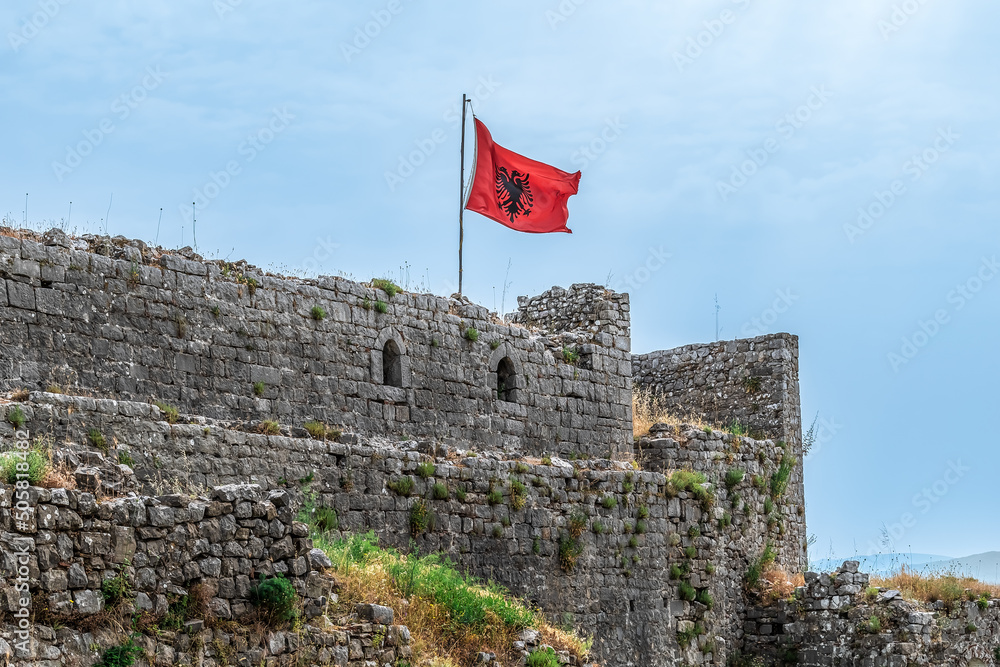 Φωτογραφία The Albanian flag flies over the ruins of Rozafa Castle against a blue sky on a summer day, Albania
