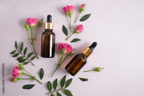 top view of two glass bottles with cosmetics, face serum against the background of pink rose buds and green leaves.