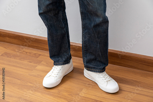Close up of man leg's with white sneakers and blue jean standing on wooden floor.