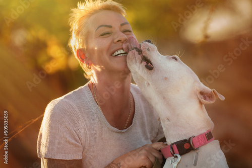 Smiling woman with white Dogo Argentino dog puppy, trying to pose, but the dog is licking and chewing her nose