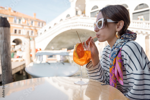 Young woman enjoying cocktail drink while sitting at outdoor cafe on the background of famous Rialto bridge and Grand Canal in Venice. Happy leisure time and vacations traveling Italy