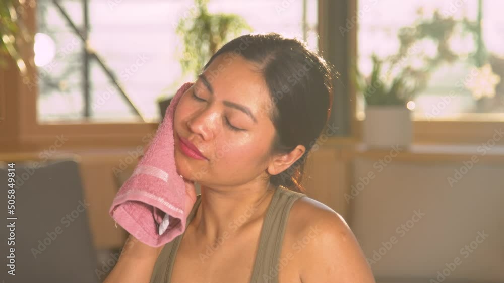 CLOSE UP: Pretty Asian woman wiping sweat from her face after intensive training. Young female person using pink towel to wipe sweat after physical exercise. Taking break during home sport activity. CLOSE UP: Pretty Asian woman wiping sweat from her face after intensive training. Young female person using pink towel to wipe sweat after physical exercise. Taking break during home sport activity.