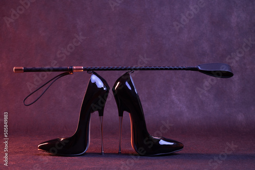 Fototapet Black shiny high heel shoes and a whip on a dark background