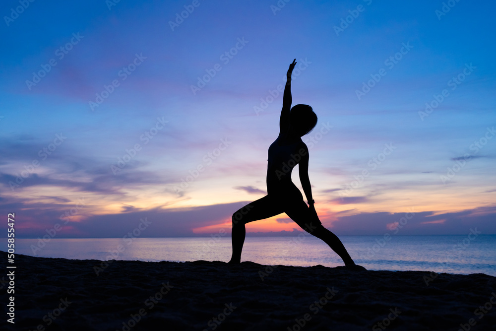 Silhouette of a beautiful woman practicing yoga asana on the beach with sunrise on twilight blue vibrant sky and calm sea in background. Peaceful Warrior, Stretching, June 21, International Yoga Day.