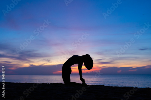 Silhouette of a beautiful woman practicing yoga asana on the beach with sunrise on twilight blue vibrant sky and calm sea in background. Camel pose, Ustrasana, June 21, International Yoga Day, Concept