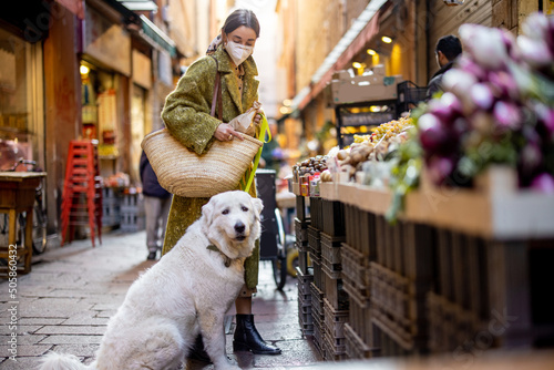 Woman in medical mask buying fruits and vegetables with dog at market stall on the famous gastronomical street in Bologna. Concept of buying local products during pandemic. Idea of Italian lifestyle