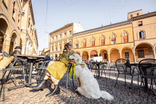 Stylish woman sitting with her white dog at outdoor cafe in the old town of Bologna city. Italian measured lifestyle and street fashion concept. Idea of traveling Italy