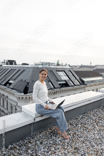 cheerful woman in jeans sitting on rooftop with laptop and smiling at camera