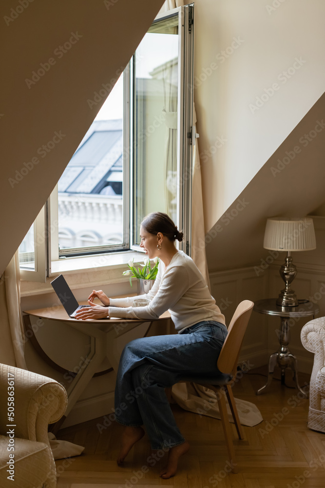 side view of woman typing on laptop in cozy attic room near window side view of woman typing on laptop in cozy attic room near window