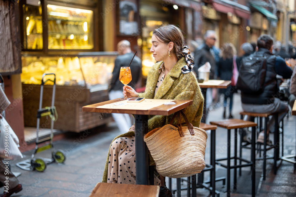 Woman sitting with a cocktail at outdoor bar or restaurant on the crowded street in Bologna. Concept of Italian lifestyle and gastronomy