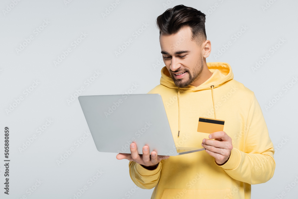 pleased man in yellow hoodie holding credit card near laptop isolated on grey. pleased man in yellow hoodie holding credit card near laptop isolated on grey.