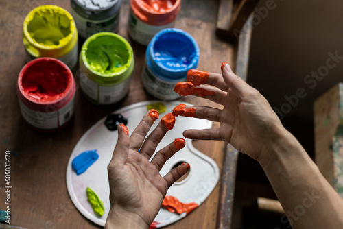 Close-up of the hands of an unrecognizable Latin American artist showing the fingers of her hand with paint, background pots of paint of various colors and palette. Concept of artist and hobby