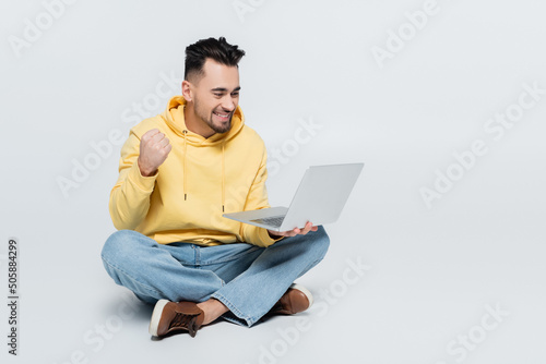 excited bookmaker showing success gesture near laptop while sitting on grey.