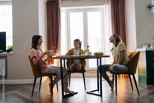 Lesbian family with their daughter sitting at the kitchen table, having breakfast and smiling © NADIN
