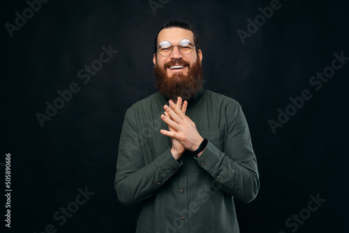 Bearded young man is holding hands together while smiling at the camera. Studio shot over black background.