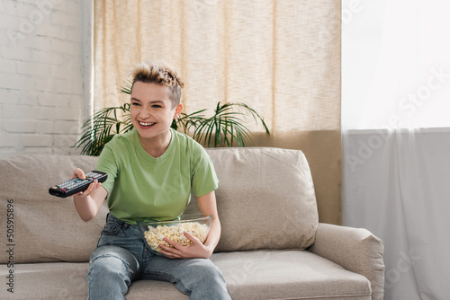 smiling pensexual person with bowl of popcorn clicking tv channels on couch at home.