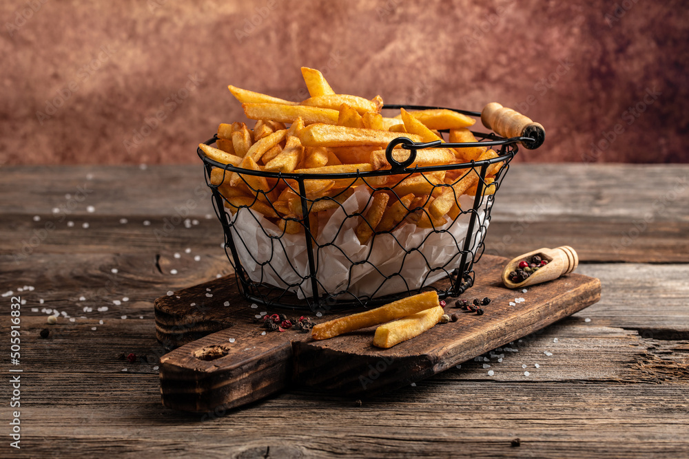 Fototapeta premium Fresh hot french fries with salt and spices on cutting board on wooden table background, unhealthy calories meal