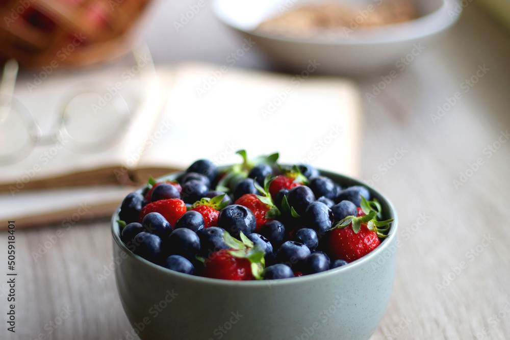 Bowl of strawberries and blueberries, open books with reading glasses, plate of chocolate chip cookies and basket of apples one the table. Selective focus. Bowl of strawberries and blueberries, open books with reading glasses, plate of chocolate chip cookies and basket of apples one the table. Selective focus.