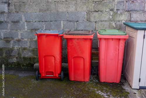 Red small wheelie recycling plastic bins in a back yard of a house by a wall. Waste disposal industry.
