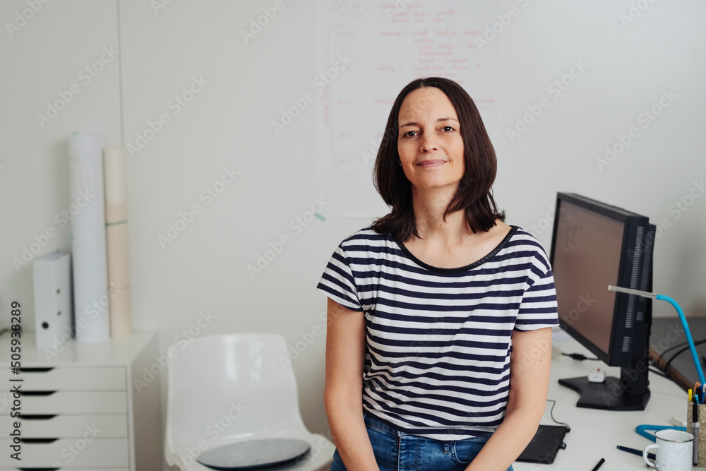 Business woman sitting on desk in office looking at camera