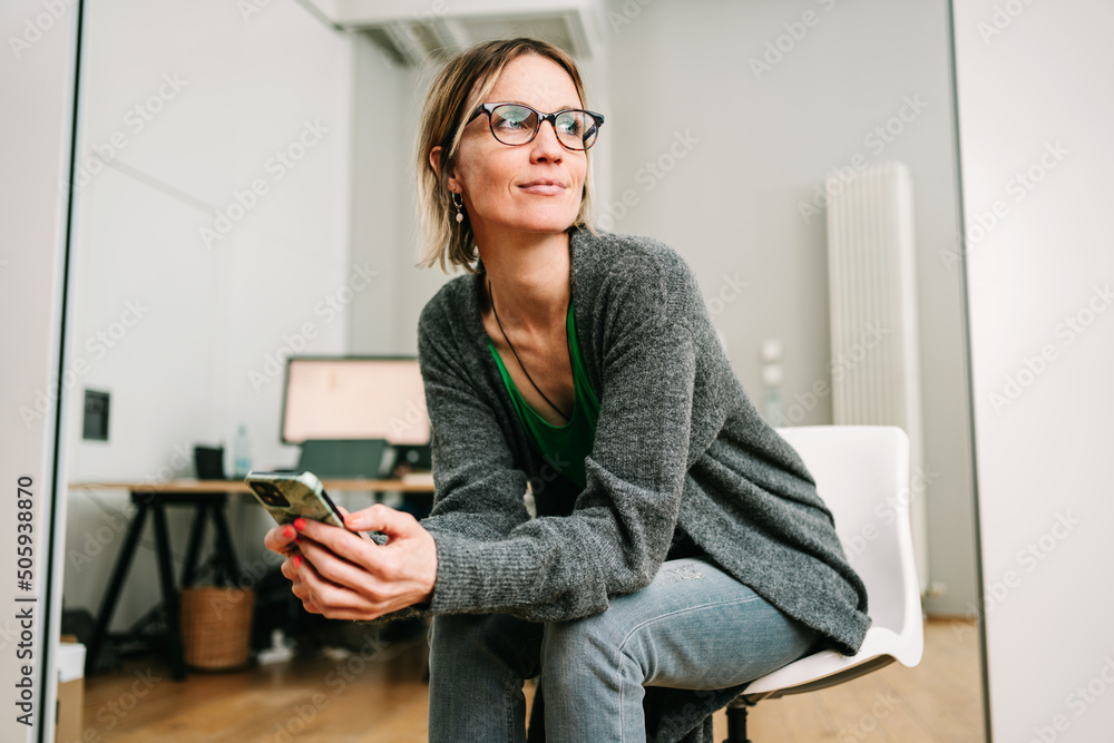 Businesswoman with glasses sits in office with a cell phone in her hand and looks thoughtfully to the side Businesswoman with glasses sits in office with a cell phone in her hand and looks thoughtfully to the side