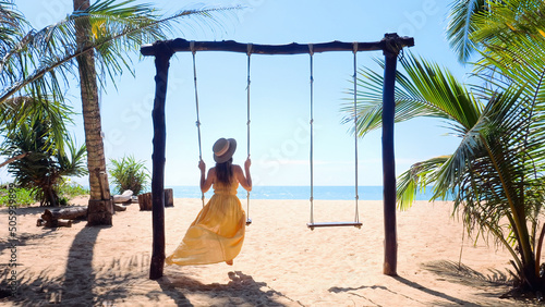 Back view of travel woman in hat sitting on swing on the beach with palm trees and turquoise sea on background. Summer vacation, tropical holidays photo