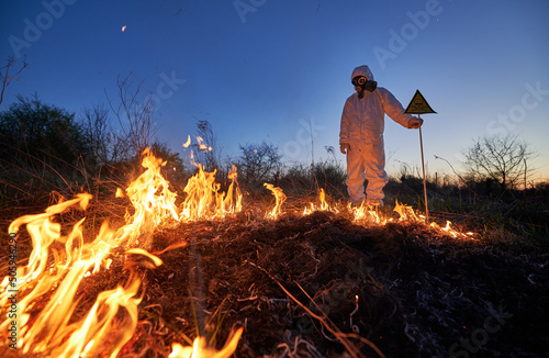 Fireman ecologist fighting fire in field at night. Man in protective radiation suit and gas mask near burning grass with smoke, holding yellow triangle with skull and crossbones warning sign.