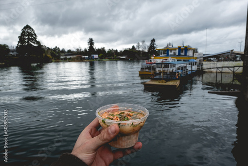Hand holding a little 'ceviche' (South American dish of fresh raw fish cured in citrus juices and spiced with chili peppers) of salmon and the river with boats and cloudy sky, Valdivia,Chile